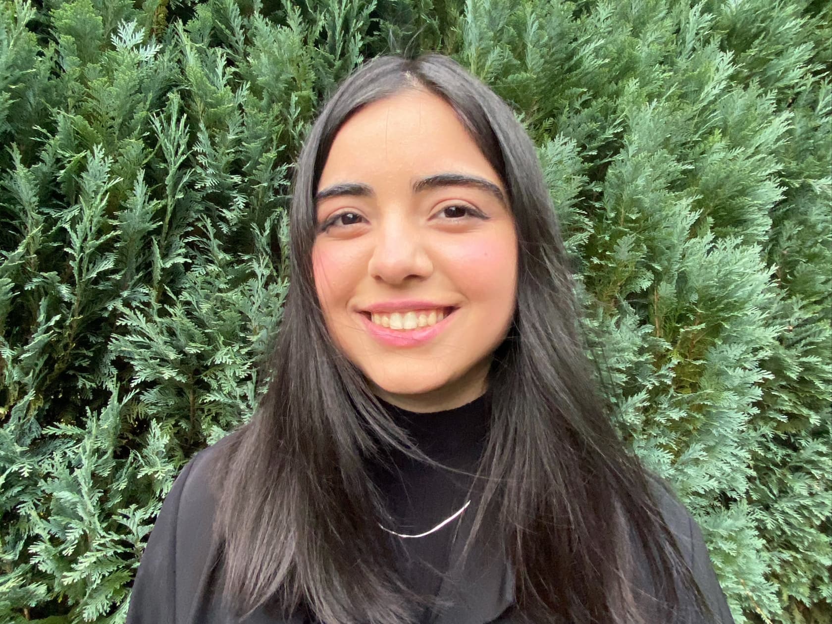 Im Zentrum des Bildes ist Nursima zu sehen wie sie freundlich lächelt. Sie trägt schwarze Kleidung und eine silberne Kette. Ihre Haare wehen im Wind. Das Foto wurde draußen geschossen. Im Hintergrund ist ein grüner Nadelbaum Strauch zu sehen.