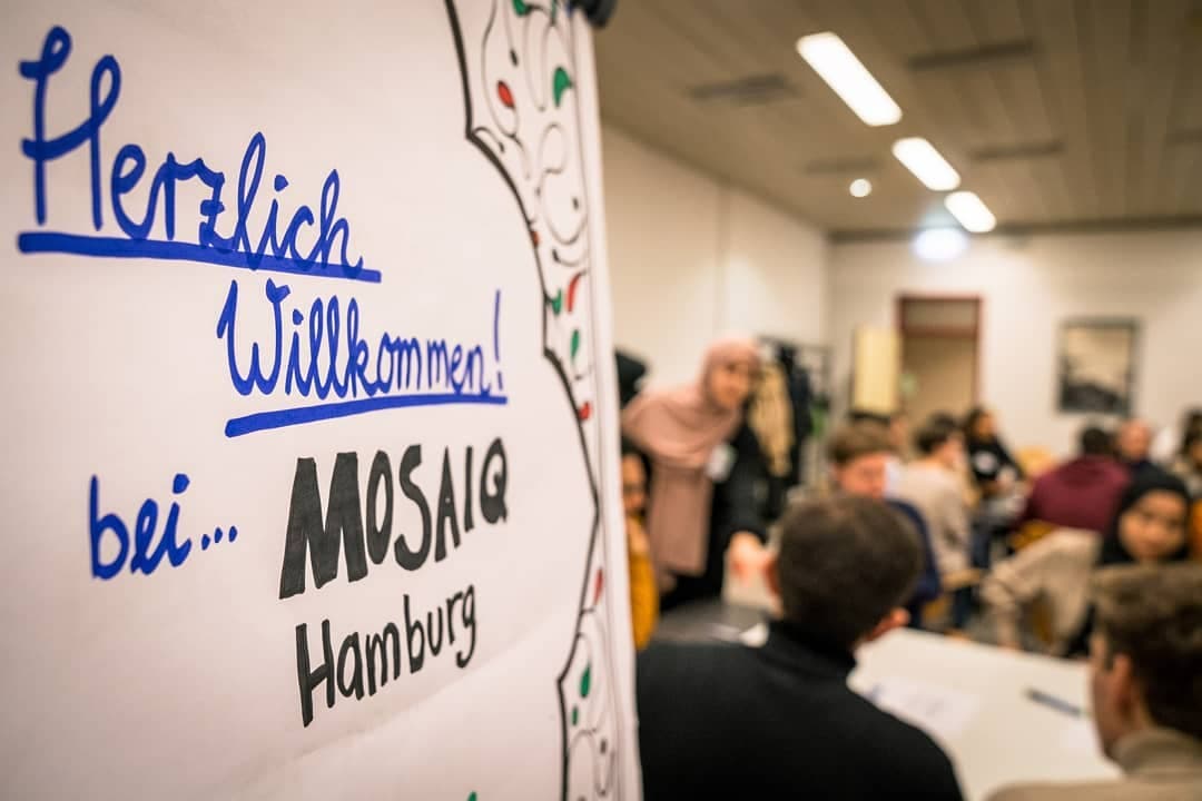 Auf der linken Seite ist ein Flipchart-Papier zu sehen, auf welchem geschmückt "Herzlich Willkommen bei Mosaiq Hamburg" steht. Auf der Rechten Seite ist ein Raum zu sehen, in verschwommener Optik, auf dem mehrere Personen miteinander reden.