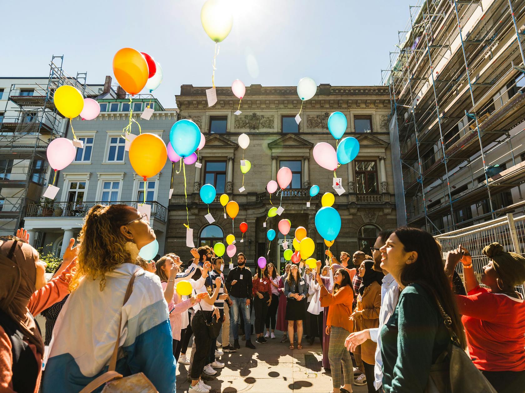Auf dem Foto ist eine Gruppe junger Menschen abgebildet, die vor einem Gebäude in einem Halbkreis stehen. Die Personen halten alle bunte Luftballons an Schnuren in der Hand und sind dabei, diese in die Luft fliegen zu lassen.
